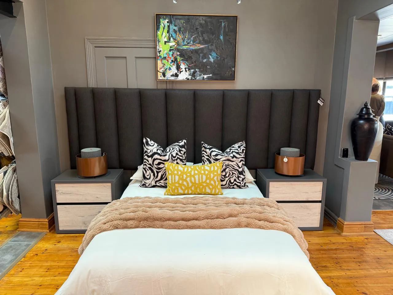 Showroom bedroom display with a charcoal upholstered headboard, patterned cushions, bedside tables and abstract artwork at Free State Curtain Parlour in Bloemfontein